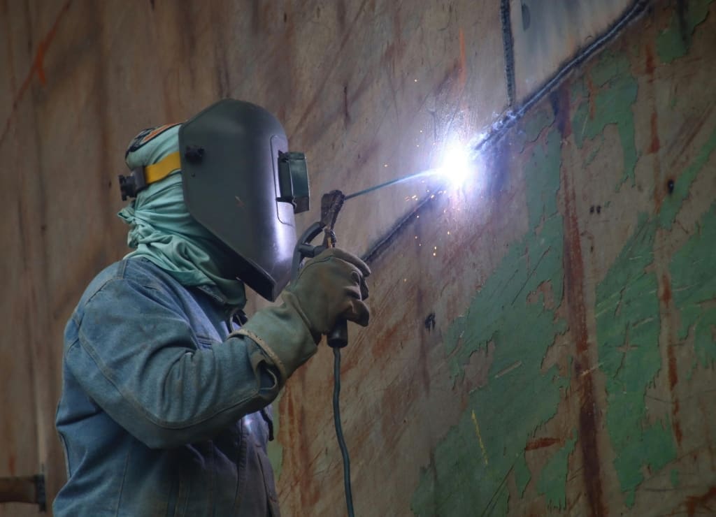 A welder in helmet and gloves running a seam on a ship’s hull plating, the arc the brightest thing in the frame, old paint and steel around the weld