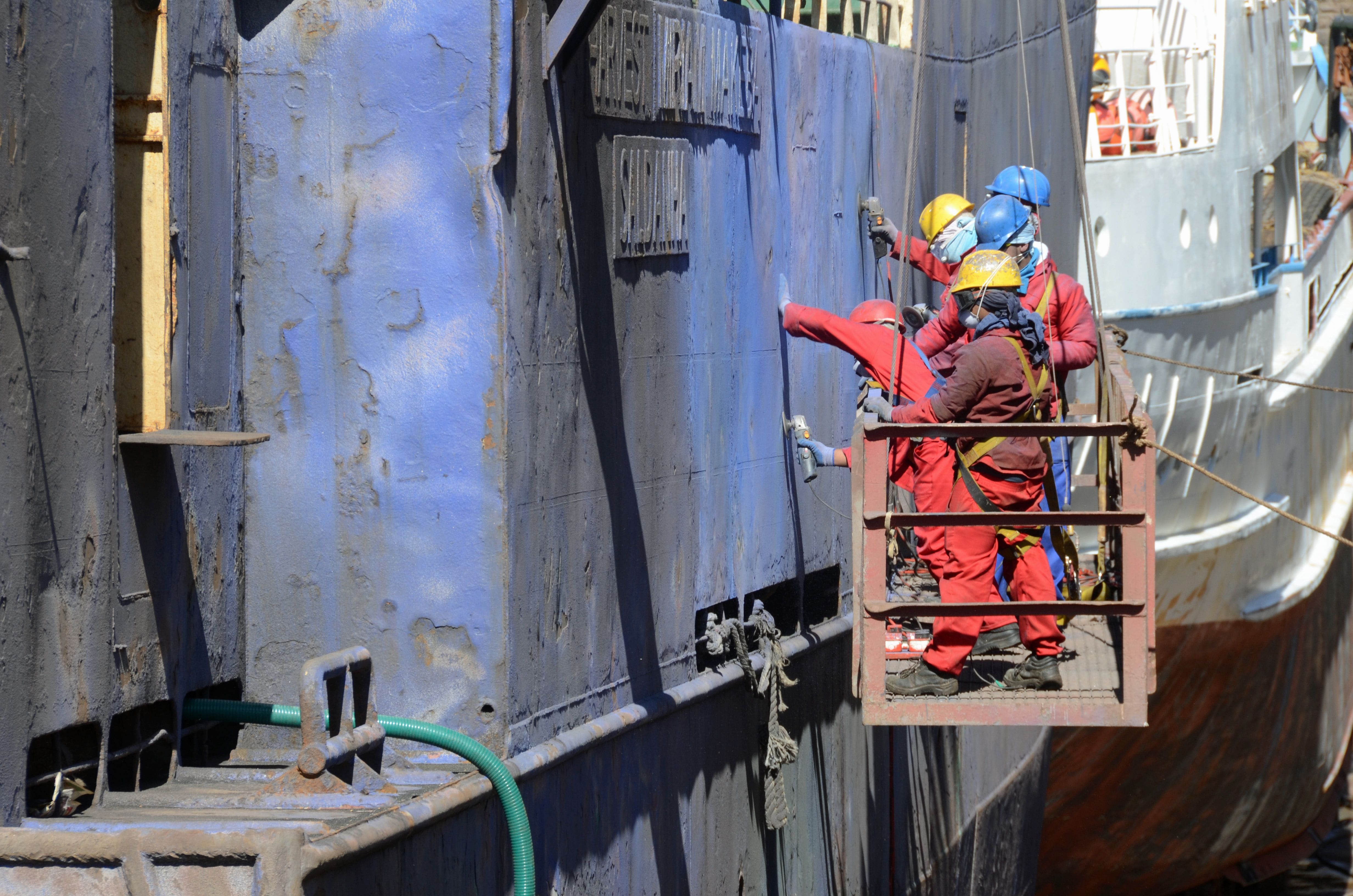 A repair crew on a suspended work platform against a ship’s hull, working on the side plating in harnesses and hard hats, patch plates and weld seams visible around them