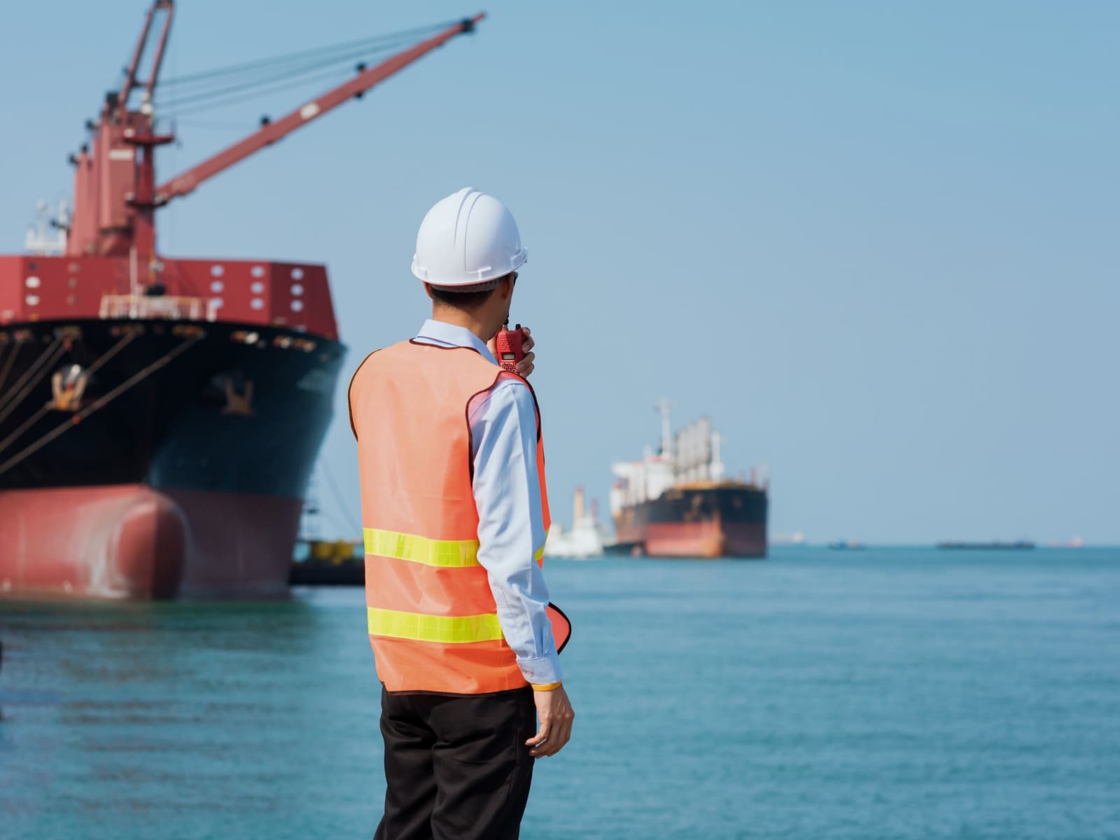 A superintendent in a hi-vis vest and hard hat on a quay, radio in hand, watching a bulk carrier lying alongside